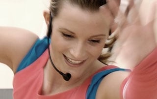 Aerobictrainerin mit Headset gibt einen Kurs, hebt die Arme in die hoch und lächelt freundlich